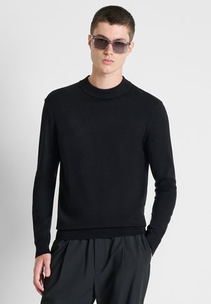 Junger Mann mit dunkler Sonnenbrille, einem schwarzen Langarmpullover und dunklen Hosen, mit einer Hand in der Tasche vor einem schlichten Hintergrund.