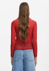 Personne aux longs cheveux bruns ondulés portant un haut rouge texturé à manches longues et un jean en denim bleu clair taille haute, vue de dos.