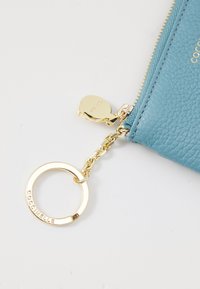Blue textured leather pouch with a gold zipper, attached gold chain, and round keyring engraved with "COCCINELLE" lettering.