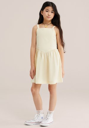 Ragazza con lunghi capelli scuri che indossa un abito senza maniche giallo pallido, calzini bianchi di pizzo, scarpe da ginnastica alte e una collana di fiori, in piedi su uno sfondo neutro.