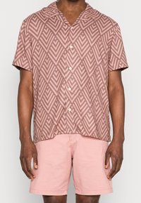 Man med kortärmad knäppskjorta i brun geometrisk mönstring och ljusrosa shorts, stående mot en enfärgad bakgrund.