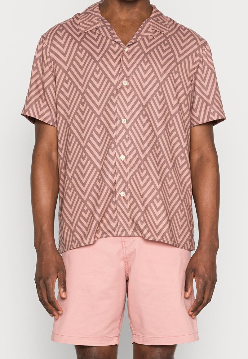 Man med kortärmad knäppskjorta i brun geometrisk mönstring och ljusrosa shorts, stående mot en enfärgad bakgrund.