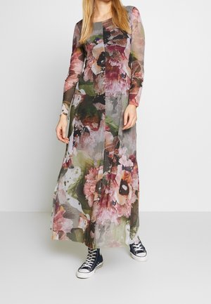 Kobieta w długiej przezroczystej, kwiecistej sukni z długimi rękawami, w czarnych płóciennych tenisówkach, stojąca na tle białego tła.