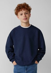 Felpa blu navy con collo rotondo, polsini a costine e taglio rilassato. Indossata sopra una camicia bianca, abbinata a jeans azzurri chiari.