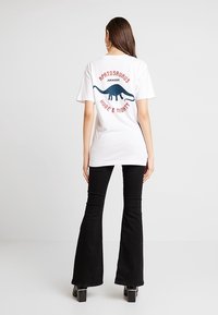 Valkoinen t-paita, jossa on sininen Apatosaurus-kuvio ja punainen teksti "JURASSIC HUGE & MIGHTY" selässä. Yhdistetty mustiin leveälahkeisiin housuihin.