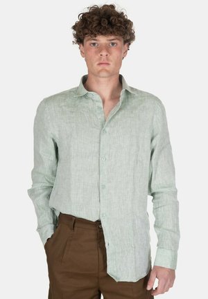 Giovane uomo con capelli ricci indossa una camicia verde chiaro con bottoni e pantaloni marroni, in piedi con una mano in tasca contro uno sfondo semplice.