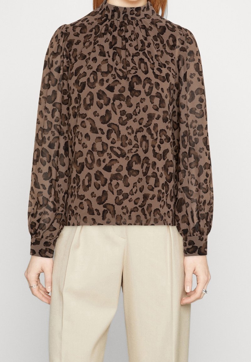 Femme portant un chemisier marron imprimé léopard à manches longues avec un col haut et un pantalon plissé beige taille haute.