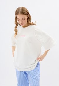 Witte oversized t-shirt met een ronde hals, voorzien van roze tekst "MIRONS" op de borst, gecombineerd met blauwe gestreepte broek. Soepele textuur.