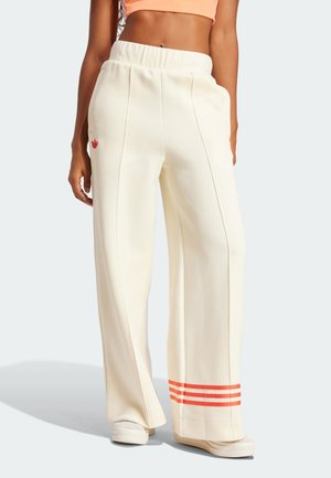 Mujer con pantalones crema de pierna ancha con tres rayas naranjas cerca del dobladillo y un pequeño logo rojo en el lado izquierdo, combinados con zapatos blancos.