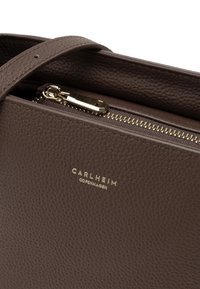 Brown leather handbag featuring a textured surface, a gold zipper, and a metal brand logo, with a simple and functional design.
