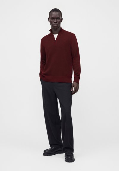 Homme debout portant un pull bordeaux à manches longues avec fermeture éclair au col, pantalon noir et chaussures noires sur un fond clair uni.