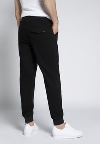 Pantaloni jogger neri a gamba affusolata con polsini elasticizzati, tasche laterali e posteriori, e un piccolo dettaglio arancione sulla tasca posteriore. Indossati con sneaker bianche.