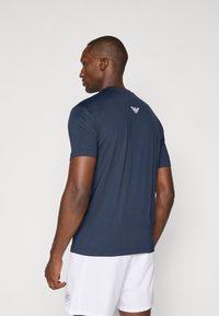 Θαλάσσιο μπλε αθλητικό μπλουζάκι κοντομάνικο με λείο υφή, στρογγυλή λαιμόκοψη και διακριτικό λογότυπο στο πίσω μέρος. Συνδυασμένο με άσπρα σορτς.