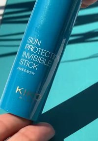 Solbeskyttende usynlig stift i en skinnende turkise tube. Har hvit tekst som merker den for ansikt og kropp. Gylden logo nederst.