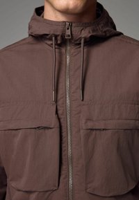 Chaqueta ligera marrón con cremallera frontal completa, dos bolsillos en el pecho, capucha ajustable con cordones y detalles de costura sutiles.