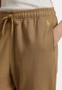 Brown sweatpants featuring an elastic waistband with a drawstring, side pockets, and a small yellow embroidered logo on the side.