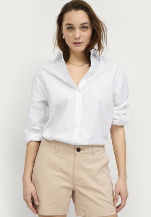 Kobieta o brązowych włosach do ramion ubrana jest w białą, zapinaną na guziki koszulę z podwiniętymi rękawami oraz beżowe spodenki, stoi na jednolitym tle.