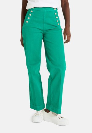 Pantalons en coton verts à taille haute, dotés de boutons sur les côtés, de jambes larges et de poignets retroussés. Associés à des baskets blanches.