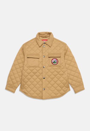 Veste matelassée marron clair avec deux poches poitrine, boutons-pression et un patch rond avec une montagne et du texte sur la poche droite.