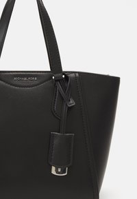 Czarna, teksturowana skórzana torba typu tote Michael Kors z podwójnymi uchwytami i wiszącą skórzaną metką z srebrnymi okuciami na białym tle.