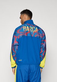 Modrá bunda sportovního soupravy s vzorovaným zadním dílem, červenými akcenty, žlutými rukávy a zlatým nápisem "BARCA". Texturovaný, lehký materiál.