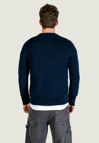 Maglione blu navy con orlo e polsini a costine, caratterizzato da una scollatura tonda e una texture liscia. Indossato sopra a una camicia bianca, presenta una vestibilità casual.