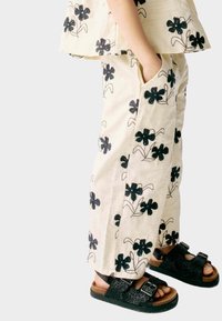 Vêtement beige clair avec broderie florale noire ; haut ample à manches courtes, pantalon assorti avec poches, associé à des sandales noires.