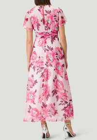Robe maxi florale rose avec de courtes manches transparentes, ornée de grands motifs de pivoine et d'une taille cintrée. Comprend un détail de dos en trou de serrure.