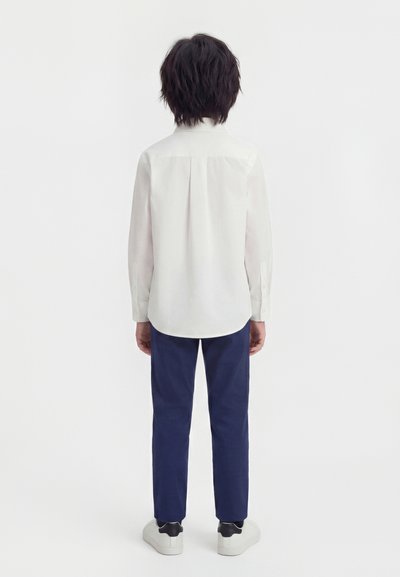 Παιδί με σκούρα μαλλιά, ντυμένο με λευκό πουκάμισο μακρυμάνικο, σκούρο μπλε παντελόνι και λευκά αθλητικά παπούτσια, στέκεται γυρισμένο προς τα πίσω σε λευκό φόντο.