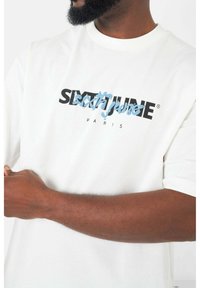 Witte katoenen t-shirt met korte mouwen, ronde hals en een grafische print in zwart en blauw met de tekst "sixtjune" en "PARIS."