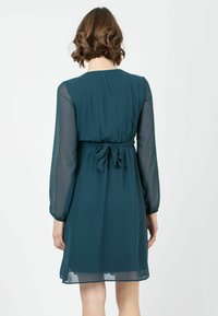 Tealfarbenes Kleid mit langen transparenten Ärmeln, tailliertem Schnitt und Schleifendetail am Rücken. Glattes Material, knielang, schlichter runder Ausschnitt.