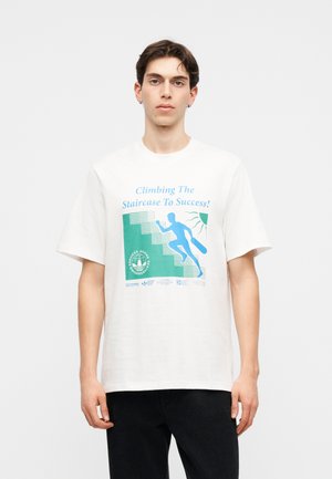 Νέος άνδρας φορώντας λευκό μπλουζάκι με μπλε γραφικό ενός ατόμου που ανεβαίνει σκάλες και με κείμενο «Climbing The Staircase To Success!»