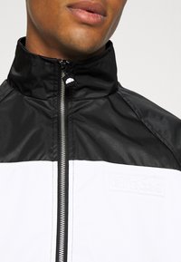 Schwarz-weißes Zip-Jackett mit hohem Kragen, glänzender Oberfläche, Logo auf der Brust und silbernem Reißverschluss. Strukturstoff mit sportlichem Design.