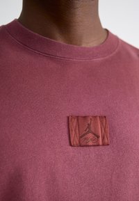 Mørkerød sweatshirt med en tekstureret brun lap med et Jordan-logo og "Fly" tekst. Crew neck-design, blød stofftekstur.