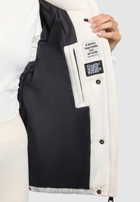 Chaqueta blanca de plumas con forro de satén azul marino y botones negros. Las etiquetas interiores incluyen instrucciones de cuidado y el logotipo de la marca en colores contrastantes.