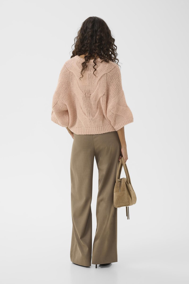 Lichtroze gebreide trui met kabelpatronen, wijde mouwen, gecombineerd met bruine high-waisted broeken en een kleine tan handtas.