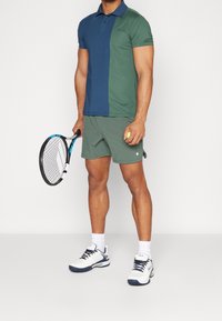 Polo de color bloque en verde y azul con cuello, combinado con pantalones cortos verdes. Sosteniendo una raqueta de tenis y una bola. Zapatos y calcetines deportivos blancos.