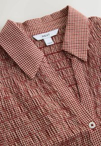 Camicia a quadri rossa e bianca con tessuto strutturato, colletto, abbottonatura frontale e dettagli arricciati lungo il corpo. Etichetta del marchio visibile.