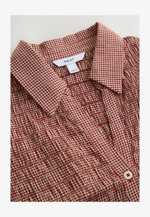 Chemise à carreaux rouges et blancs présentant un tissu texturé, un col, une patte de boutonnage à l'avant et des détails froncés le long du corps. Étiquette de la marque visible.