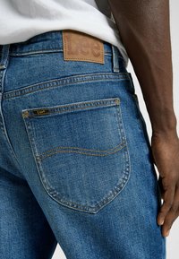 Jeans de mezclilla azul con un parche de cuero de la marca Lee en la cinturilla trasera. Presenta un diseño clásico de bolsillos y detalles de costura en color marrón.
