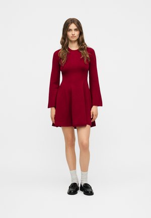 Γυναίκα με μακριά καστανά μαλλιά, που φοράει μακρυμάνικο μαρόν φόρεμα σε γραμμή Α, λευκές κάλτσες και μαύρα loafers, στέκεται σε απλό λευκό φόντο.