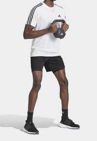 Camiseta atlética blanca con diseño de tres rayas negras, combinada con pantalones cortos negros. La persona sostiene un kettlebell negro, vistiendo zapatos y calcetines negros.