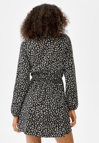 Kvinna med lockigt hår som bär en svart långärmad klänning med ett vitt blommönster, sedd bakifrån.