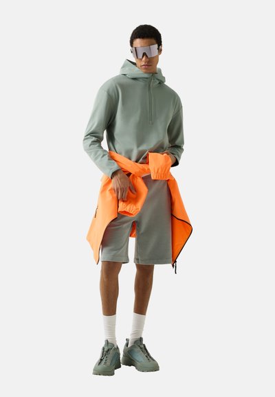 Homme portant un sweat à capuche vert, un short assorti, des baskets vertes, des chaussettes blanches et des lunettes de soleil réfléchissantes, avec une veste orange attachée autour de la taille.
