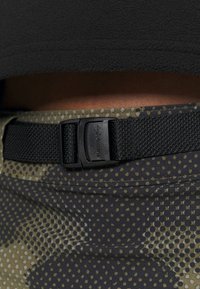 Cinturón negro con hebilla de plástico, abrochado a unos pantalones estampados en verde oscuro y negro con diseños circulares y tela texturizada.