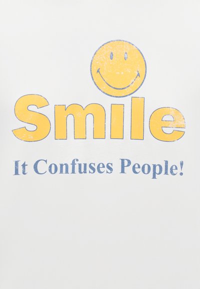 Λευκό πουκάμισο με κίτρινο γραφικό με πρόσωπο που χαμογελά πάνω από το κείμενο «Χαμογέλα» σε κίτρινο και «Μπερδεύει τους ανθρώπους!» σε μπλε. Σχέδιο με εφέ φθοράς.