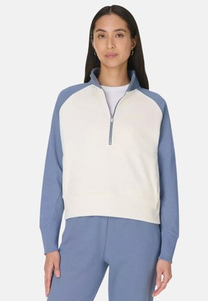 Vrouw draagt een wit-blauwe trui met een kwartritssluiting en blauwe broek, staand met een neutrale uitdrukking tegen een effen achtergrond.