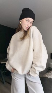 Mikina v světle béžové látce s oversize střihem a žebrovaným výstřihem, spárovaná se šedými tepláky. Černá čepice a zlaté náušnice dodávají detail.