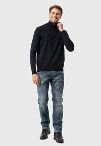 Marineblauer Strick-Reißverschluss-Pullover mit geripptem Kragen, kombiniert mit ausgewaschenen blauen Jeans und schwarzen Schnürstiefeln. Das Model steht in entspannter Haltung.