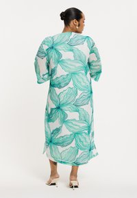 Doorzichtig, lang kledingstuk met een bloemenpatroon in tinten van teal en groen op een witte achtergrond. Kenmerkt zich door brede mouwen en splitten aan de zijkanten voor gemakkelijk bewegen.
